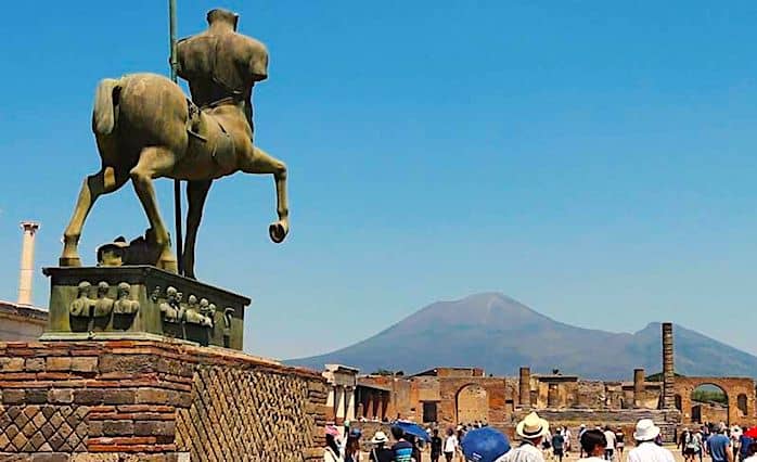People walking around the Forum of Pompeii with various Roman temples and Mt. Vesuvius in the background.